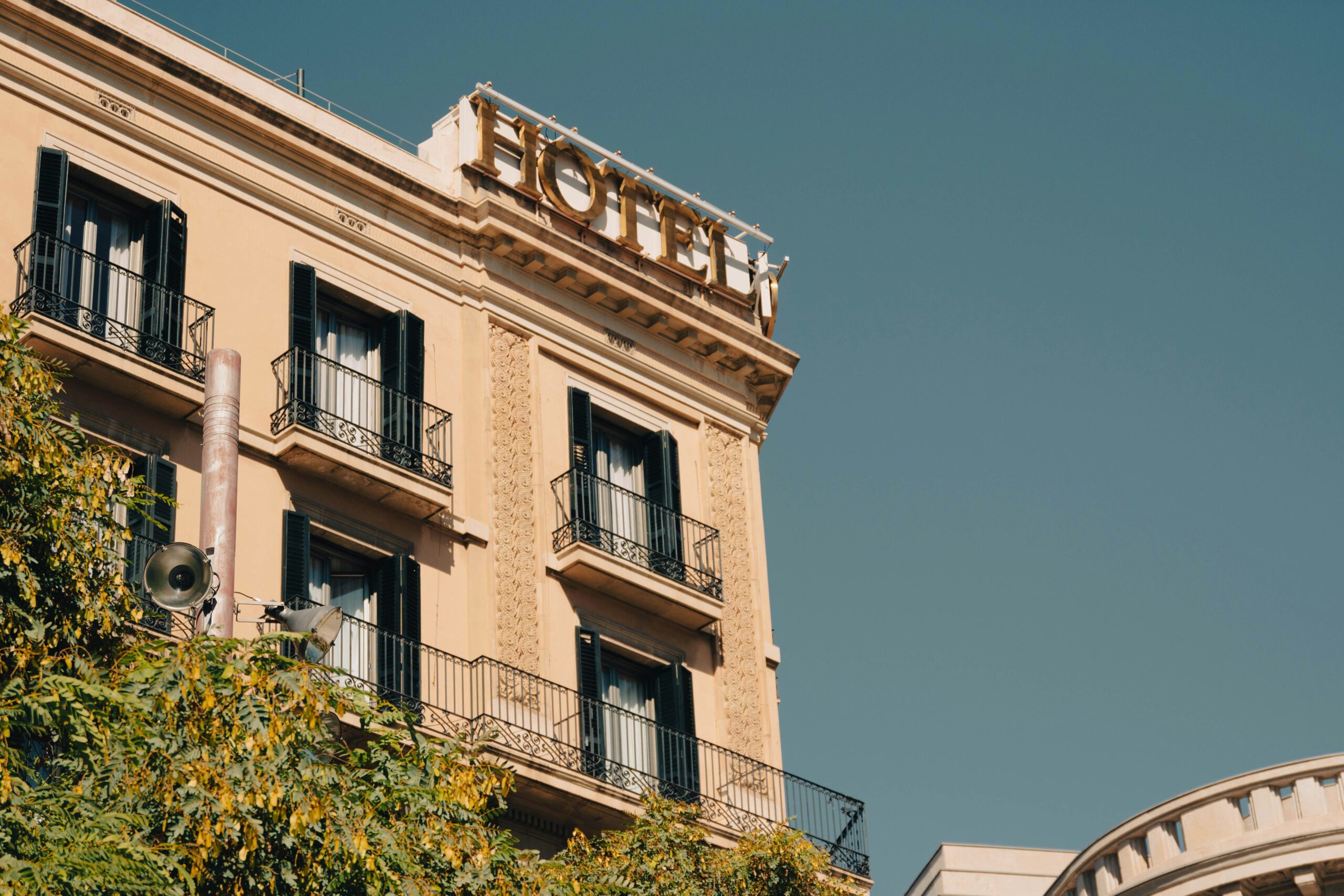 ana-lanza-aroQQ_iz0Jg-unsplash A close-up view of the upper floors of a beige European-style building with ornate architectural details, balconies with dark shutters and wrought-iron railings, and a rooftop sign reading “HOTEL.”